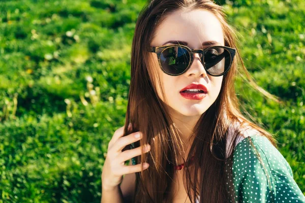 Giovane bella ragazza con i capelli scuri, indossa occhiali da sole e con il rossetto rosso sulle labbra seduto su erba verde — Foto Stock