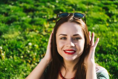 güzel genç gülümseyen kız kırmızı ruj dudakları ve güneş gözlüğü ile yeşil çayır üzerinde oturur