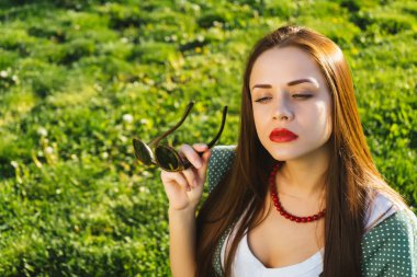 güzel genç gülümseyen kız kırmızı ruj dudakları ve güneş gözlüğü ile yeşil çayır üzerinde oturur