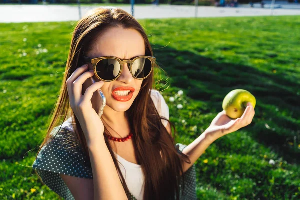 Joven enojada con gafas de sol hablando por teléfono y sosteniendo una manzana en su mano — Foto de Stock
