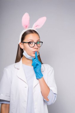 genç bir kız tıbbi Beyaz önlük ve yüz maskesi, gözlük ve RIM tavşan kulakları şeklinde bir işaret parmağı ağzında tutar.