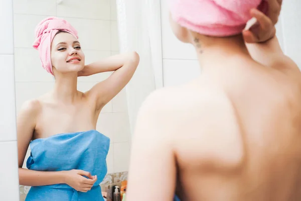 Молода красива дівчина з рожевим рушником на голові стоїть біля дзеркала і дивиться на себе, посміхаючись — стокове фото