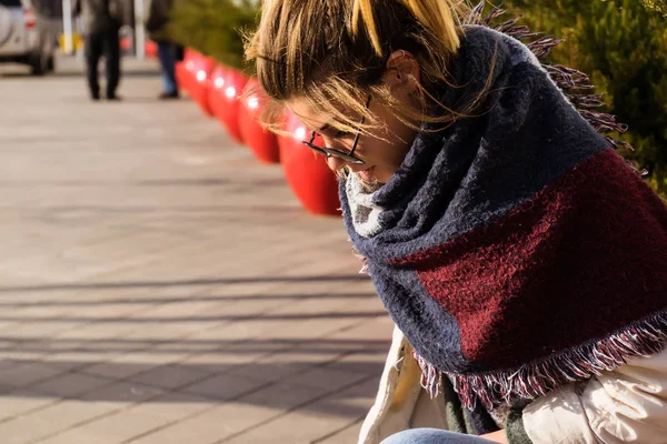 Young beautiful girl in sunglasses and fashionable scarf sits on the street and looks down — Stock Photo, Image