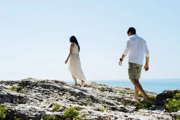 Un ragazzo e una ragazza camminano da soli lungo le rocce sullo sfondo dell'oceano, sagome irriconoscibili dal retro — Foto Stock