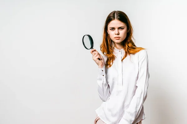 Linda menina ruiva em uma camisa branca grande está segurando uma lupa e olha para a câmera, isolado — Fotografia de Stock