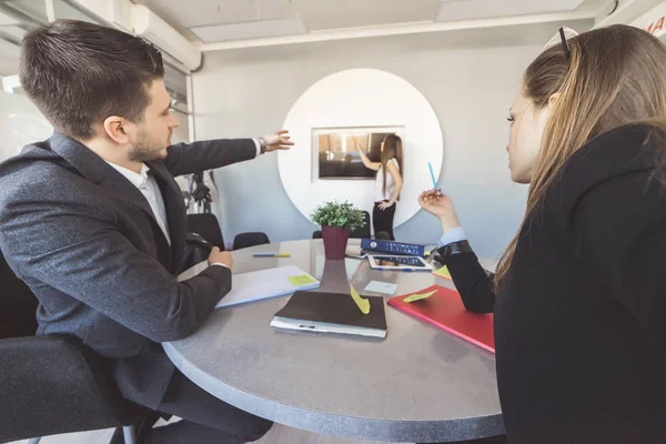 La ragazza sullo sfondo spiega le sue idee di business ai suoi colleghi e li mostra sullo schermo della TV. Giovane uomo e ragazza in primo piano seduti e puntando le mani verso la TV — Foto Stock