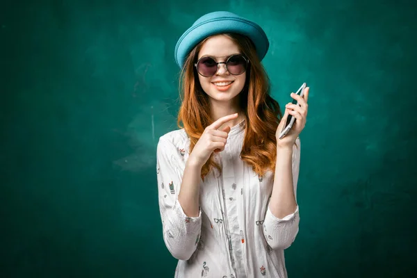 Ragazza alla moda in un cappello turchese e occhiali da sole sta tenendo un telefono tra le mani e sorridente guarda la fotocamera — Foto Stock