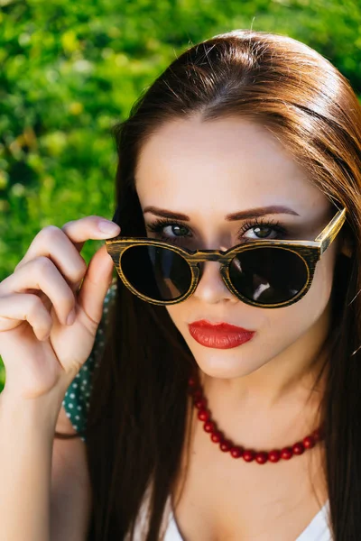 Young beautiful girl in sunglasses sits on green grass and looks at camera — Stock Photo, Image