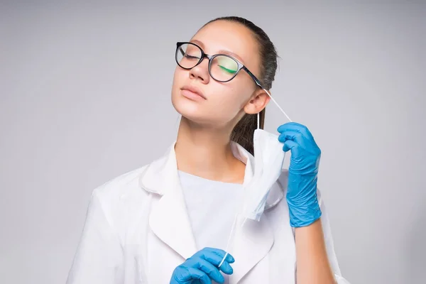 Giovane medico ragazza di successo in occhiali, vestaglia medica bianca rimuove una maschera protettiva da se stessa — Foto Stock