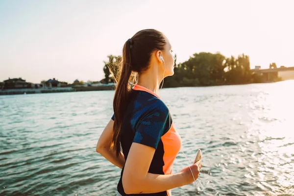 young sporty girl runs by the river at sunset, listens to music on headphones