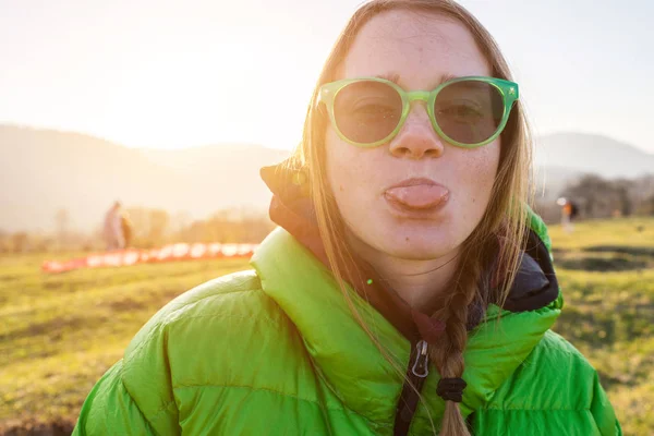 Joven chica divertida en chaqueta verde y gafas verdes se encuentra en el fondo del sol y muestra la lengua — Foto de Stock