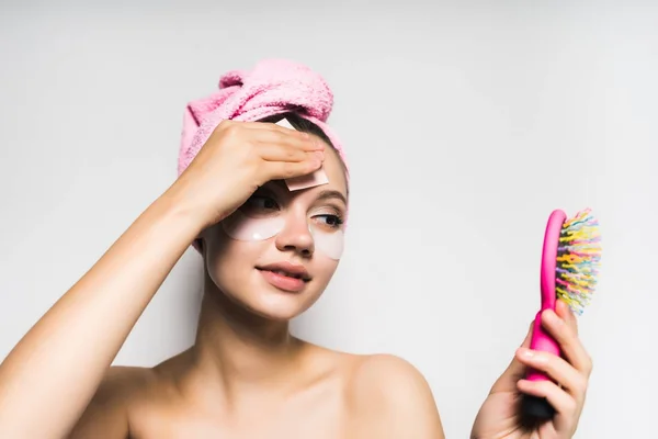 Молодая красивая девушка с розовым полотенцем на голове и силиконовыми пятнами под стеклом смотрит в зеркало — стоковое фото