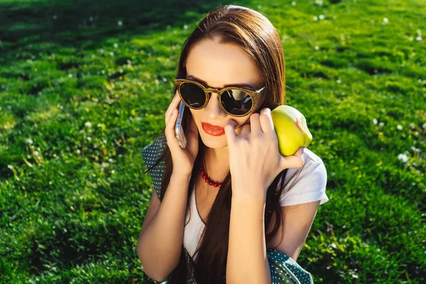 Talking woman holds mobile,sitting on grass in dress,sunglasses