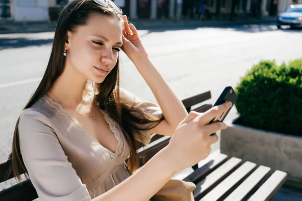 Joven hermosa chica en un vestido beige y gafas de sol se sienta en un banco de madera en la calle y sostiene en las manos su teléfono inteligente — Foto de Stock