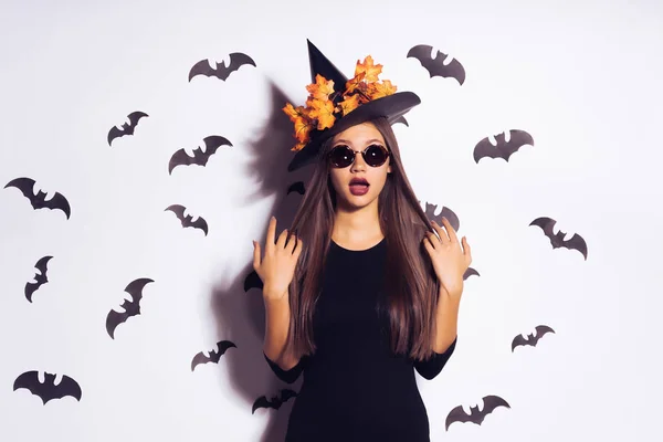 Egy divatos Helluin boszorkány egy nagy kalap levelei kör v.-védő szemüveg néz ki a kamera. a lány lesz a Halloween party. denevérek, a fehér háttér — Stock Fotó