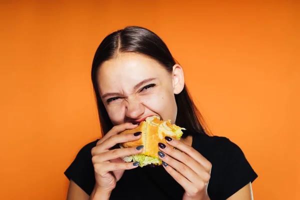 Menina ansiosamente come um hambúrguer apetitoso um fundo laranja, isolado — Fotografia de Stock