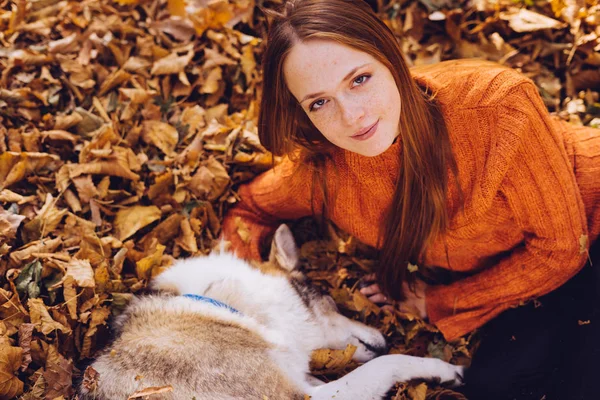 Den rödhåriga tjejen ligger i torra höstlöv med sin hund och ser in i kameran — Stockfoto