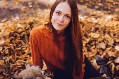 şık bir turuncu kazaklı genç güzel kızıl saçlı kız köpeğini düşen sonbahar yaprakları etrafında park ile yürüyor