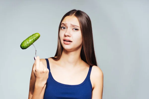 A young beautiful girl in a blue top wants to lose weight, holds a useful cucumber in her hands and looks perturbed — Stock Photo, Image