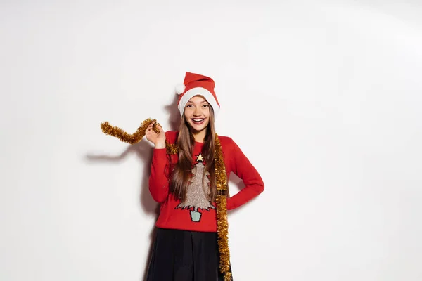 Una joven hermosa chica feliz celebra el Año Nuevo y la Navidad, con una gorra roja y un suéter caliente de moda, con un oropel dorado alrededor de su cuello —  Fotos de Stock