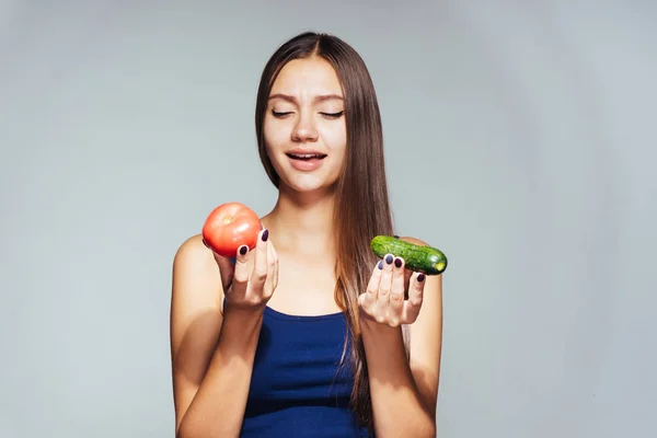 Una chica dulce joven en una parte superior azul quiere perder peso, tiene un pepino útil y un tomate en sus manos — Foto de Stock