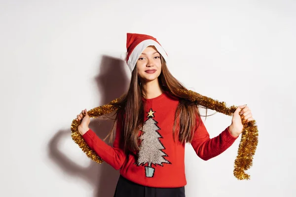 Una joven hermosa chica celebra el Año Nuevo y la Navidad, con una gorra roja y un suéter de moda, con un oropel dorado alrededor de su cuello —  Fotos de Stock