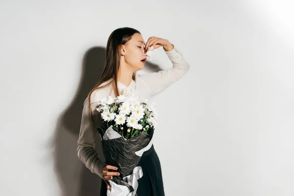 Uma jovem menina bonita em uma camisa branca está segurando um grande buquê de cet branco em suas mãos e espirra porque ela é alérgica a flores — Fotografia de Stock