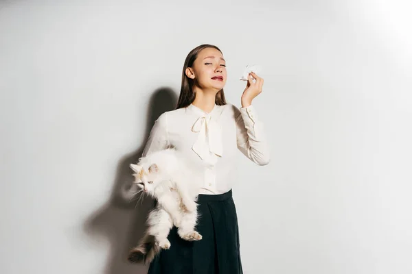 Una joven hermosa chica en una camisa blanca está sosteniendo un gran gato mullido blanco en sus manos y estornuda porque es alérgica a los animales — Foto de Stock