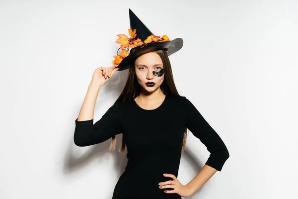 Una joven chica gótica sexy celebra halloween en la imagen de una bruja, en un gran sombrero negro, adornado con hojas amarillas de otoño, se ve confiado —  Fotos de Stock