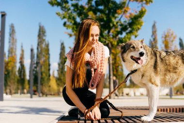 şık kazaklı genç güzel kızıl saçlı kız köpeğini, mavi gökyüzü ile parkta yürüyor