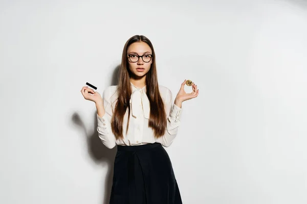 Ein junges erfolgreiches modernes Mädchen mit Brille hält einen goldenen Bicoin und eine Bankkarte in der Hand — Stockfoto