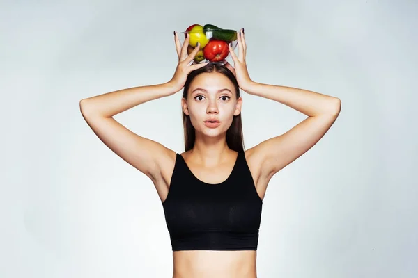 Uma jovem atlética em um top preto mantém o controle de sua figura, segura um prato com vegetais úteis e frutas em suas mãos — Fotografia de Stock
