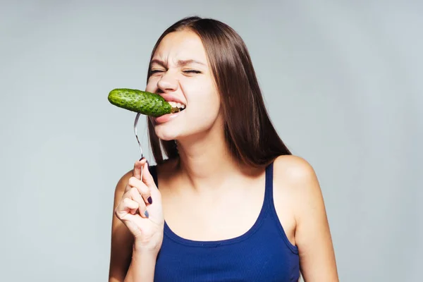 Young beautiful girl in a blue top sits on a diet, eagerly eats a low-calorie cucumber — Stock Photo, Image