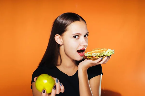 Молода красива дівчина хоче схуднути, але з нетерпінням чекає шкідливе калорійне бургер і тримає в руці корисне яблуко — стокове фото