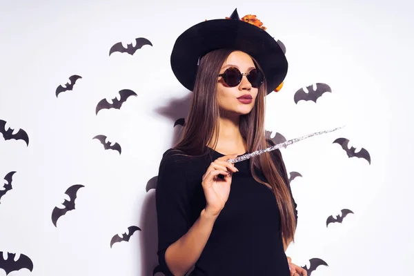 Una giovane ragazza gotica sexy allegra celebra Halloween, vestita con una strega, con un grande cappello nero, occhiali da sole, tiene una bacchetta magica — Foto Stock