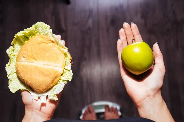 Uma jovem quer perder peso, fica na balança, segura uma maçã útil e um hambúrguer prejudicial — Fotografia de Stock