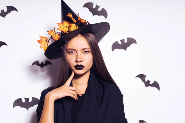 Ein junges sexy Gothic Girl feiert Halloween, gekleidet in eine Hexe, auf ihrem Kopf einen großen schwarzen Hut, der mit gelben Herbstblättern verziert ist — Stockfoto