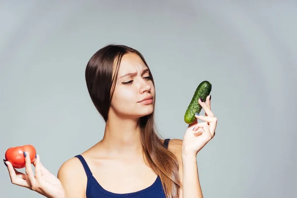 Ein junges athletisches Mädchen in blauem Top beobachtet ihre Figur, hält eine nützliche kalorienarme Gurke und eine Tomate in der Hand — Stockfoto