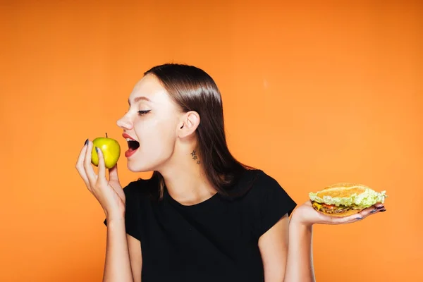 Молодая красивая девушка хочет похудеть, держит полезное яблоко и вредный калорийный бургер — стоковое фото