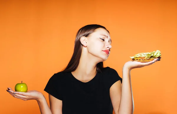 A young beautiful girl wants to lose weight, holds a useful apple and a harmful calorie burger — Stock Photo, Image