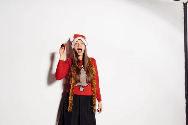 Όμορφη κοπέλα γιορτάζει την Πρωτοχρονιά και τα Χριστούγεννα, σε ένα κόκκινο σκουφάκι, χαριτωμένο πουλόβερ και χρυσά στολίδια στο λαιμό της — Φωτογραφία Αρχείου