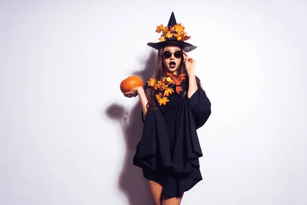 Una giovane gotica sexy gotica festeggia Halloween, a immagine di una strega, sulla sua testa un grande cappello nero decorato con foglie autunnali gialle tiene in mano una zucca — Foto Stock