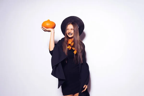 A young sexy gothic girl celebrates halloween, in the shape of a witch, in a big hat, holds an orange pumpkin — Stock Photo, Image