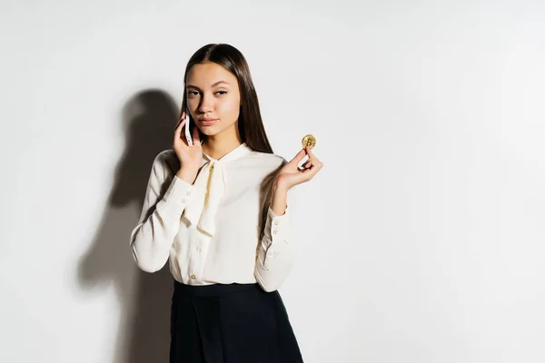 Ein junges modernes erfolgreiches Mädchen in weißem Hemd hält einen goldenen Bitcoin in der Hand und telefoniert über die Kryptowährung — Stockfoto