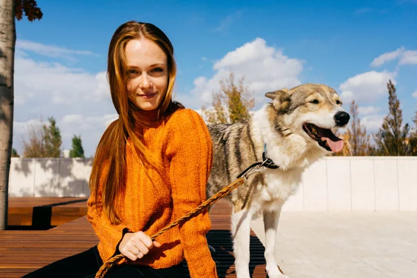 Flicka i en orange tröja sitter på en bänk med sin hund — Stockfoto