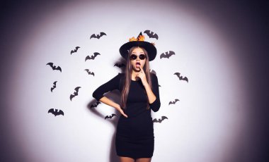 gizemli Gotik seksi kız büyük bir siyah şapka ve siyah güneş gözlüğü, parmağını ağzına koymak bir Cadılar Bayramı için bir cadı giyinmiş ve şaşırmış görünüyor