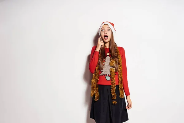 Joven feliz chica celebra Año Nuevo y Navidad, en una gorra roja y suéter, con un oropel de oro en su cuello, se ve sorprendido y entusiasta — Foto de Stock