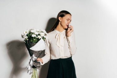 a beautiful long-haired girl in a white shirt sneezes because she is allergic to the flowers she holds in her hand