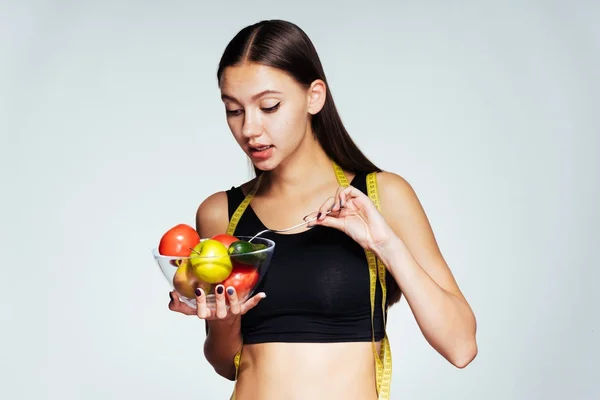 Bela menina esbelta em um top de esportes preto observa sua figura, segura um prato com vegetais úteis e frutas e um garfo — Fotografia de Stock