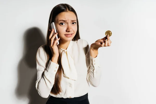 Uma bela menina de sucesso parece com raiva, fala ao telefone sobre a moeda criptomoeda e detém um bitcoin de ouro em sua mão — Fotografia de Stock